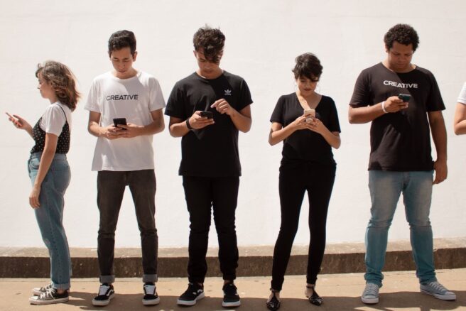 Joukko nuoria seisoo rivissä ja pelaa puhelinta.