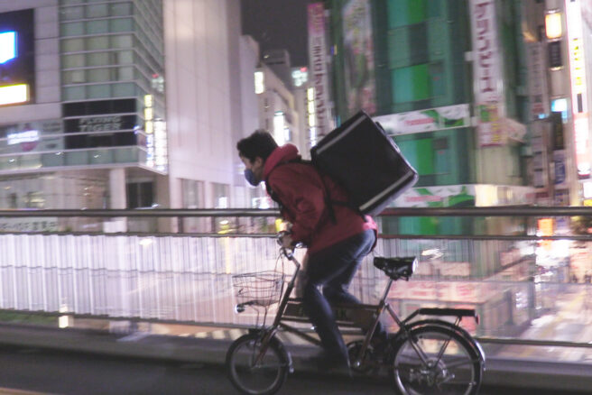 Kuvituskuva Tokyo Uber -elokuvasta. Mies pyöräilee suurkaupungin kadulla iltavaloissa.