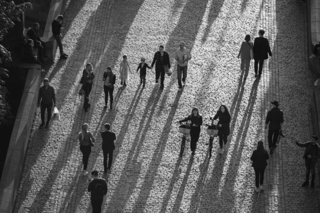 Mustavalkoinen kuva, jossa ihmisiä kadulla, aurinko heittää pitkät varjot kadulle