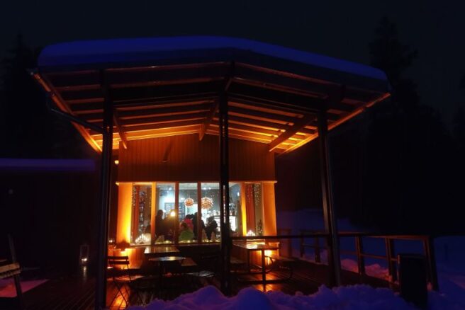 Talvinen hämärä kuva kahvilan ikkunasta sisään, pöydän ääressä on ihmisiä kokoontuneena keskustelemaan