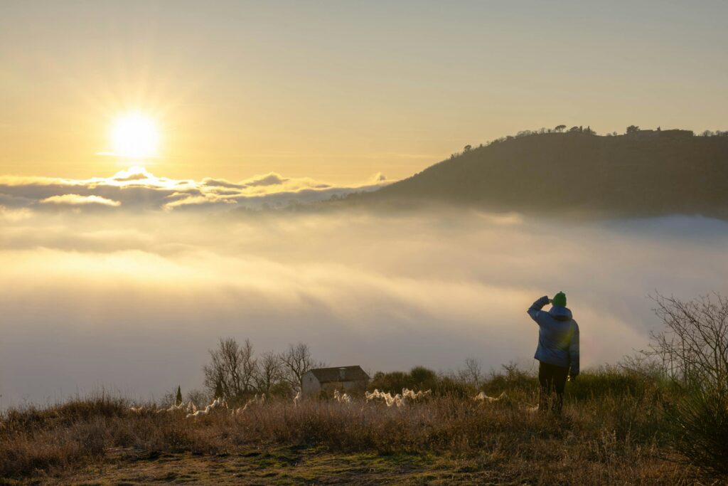 Ihminen seisomassa mäen rinteessä ihastelemassa auringonnousua pilvien takaa