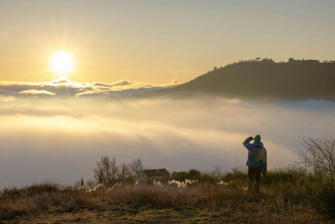 Ihminen seisomassa mäen rinteessä ihastelemassa auringonnousua pilvien takaa