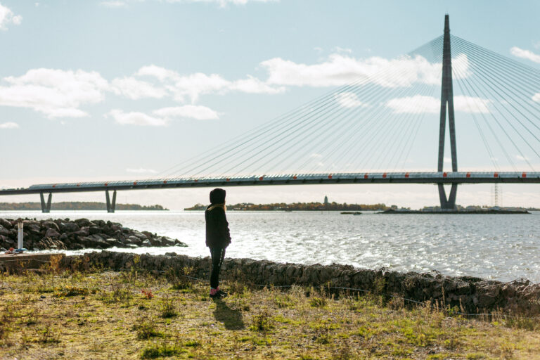 Stillkuva dokumenttielokuvasta Korvia huumaava hiljaisuus. Henkilö seisoo kevätmaisemassa, taustalla silta.